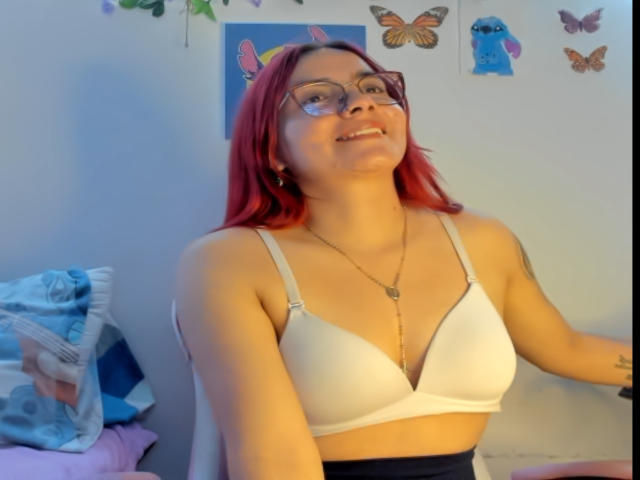 EimmyHoskX with red hair, glasses, white bra, and necklace, smiling, showing small tits, tattoo on left arm, sitting on bed with blue and pink blankets.
