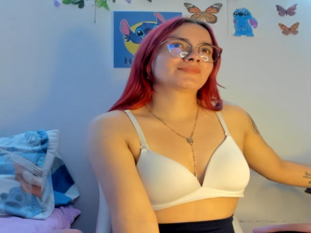 EimmyHoskX with red hair, glasses, white bra, small tits, smiling, tattoo on arm, necklace, blue and pink bed, wall art, butterflies, and owl in a softly lit room.