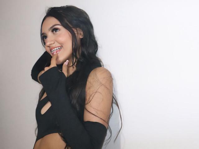 MiaaRyder with long black hair, smiling, touching her finger to her lips, wearing a black cut-out top, showing her tits and arms. White background.