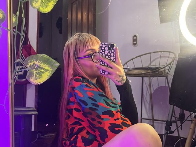 KallySpice with pink hair, glasses, and a colorful camo dress, taking a mirror selfie with her phone, showing a tattoo on her hand, and a ring on her phone case.