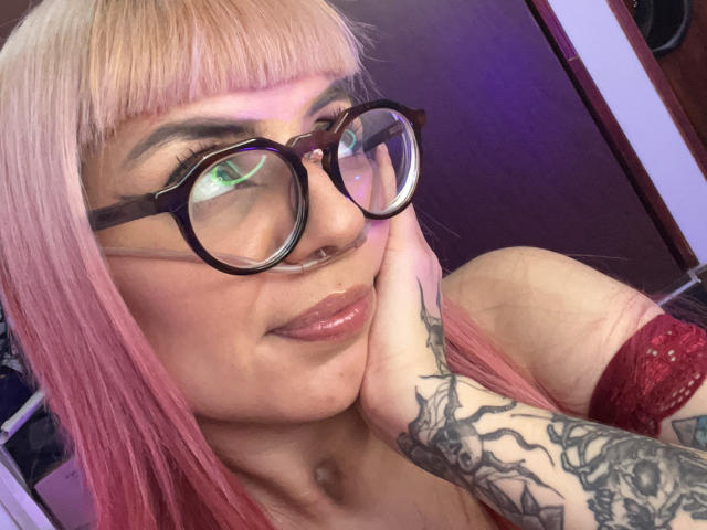 KallySpice with pink hair, glasses, nose ring, and tattooed arm, resting her chin on her hand, showing off shiny lips and a confident smile. Boobs in red lace visible.