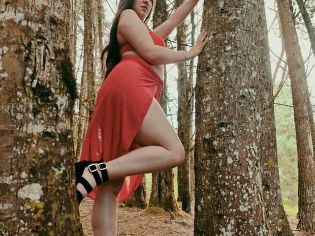 CamilaGomez with long brown hair, red dress, black-and-white shoes, standing against a tree, showing her thigh, holding the trunk, forest background.