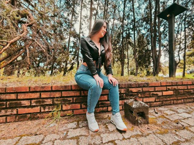 CamilaGomez assise sur un mur de briques, portant une veste en cuir noir, un jean bleu et des baskets blanches, avec de longs cheveux bruns et un sourire, dans une zone boisée..