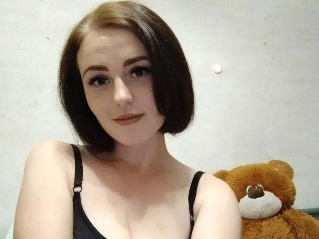 KasandraZ69 has short brown hair, black tank top, showing her tits, standing with a brown teddy bear, light skin, subtle smile, makeup, and small nose.