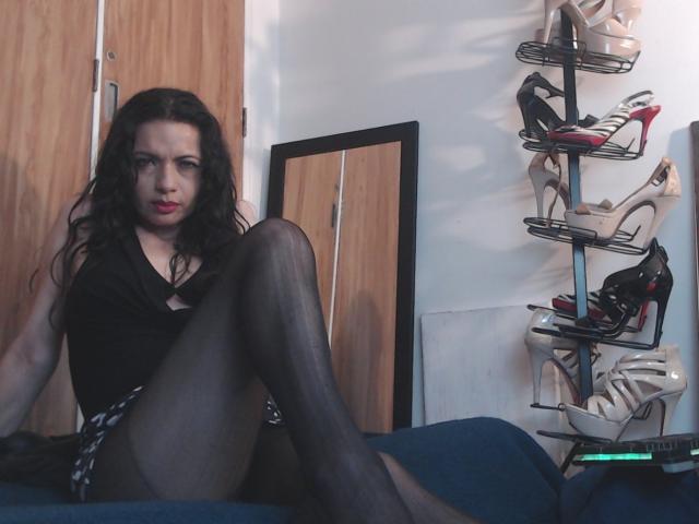 AfroditaRgenis with long black hair, wearing black top, sheer black pantyhose, and white high heels, sitting with legs up, showing her ass, in a room with shoe rack.
