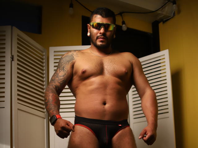 BastianGreco, muscular, tan-skinned, tattooed arm, wearing black mesh underwear with red trim, reflective sunglasses, black wristband, standing, chest hair, trimmed beard, yellow wall, white folding doors.