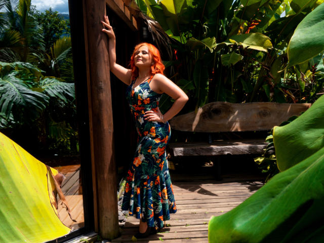 RubiQueenS with fiery red hair, wearing a floral dress, stands in a wooden hut, one arm raised, showcasing her curvy ass and tits, surrounded by lush greenery.