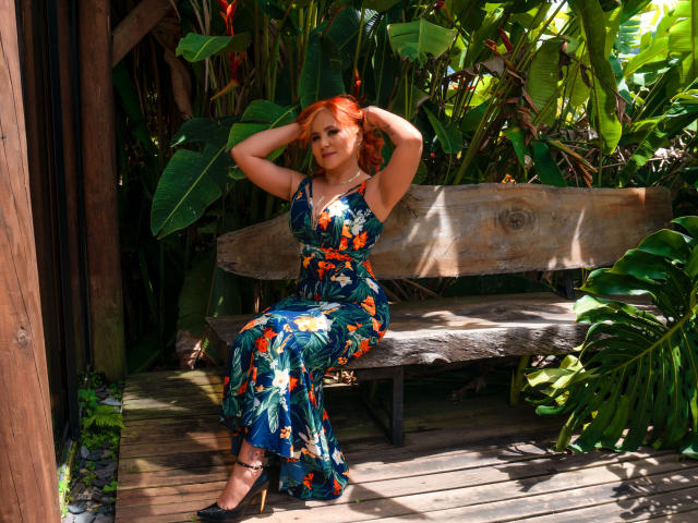 RubiQueenS with red hair, wearing a floral blue dress, sits on a wooden bench, hands behind head, black heels, lush green plants, sunny, relaxed, sexy.
