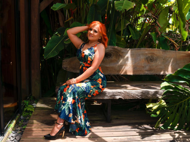 RubiQueenS with red hair, wearing a floral dress, sitting on a wooden bench, hand in hair, black heels, green leaves and wood background, sunlight, curvy, cute.