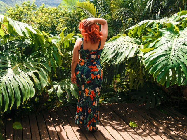 RubiQueenS with red hair, floral dress, and back to camera, hand in hair, standing on wooden path, surrounded by large green leaves, sun shining, tropical vibe.