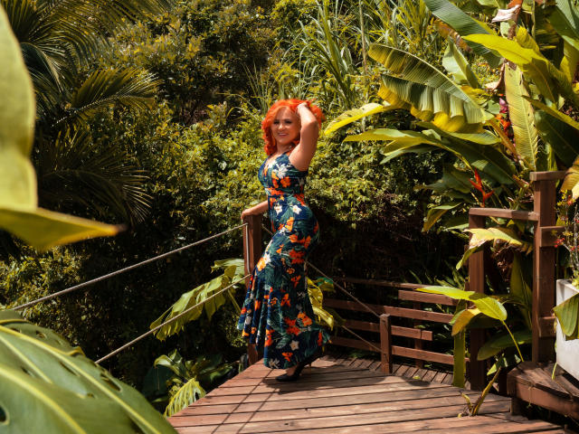 RubiQueenS with red hair, wearing a blue floral dress, standing on a wooden bridge, hand in hair, surrounded by lush green plants. Her ass is slightly visible.
