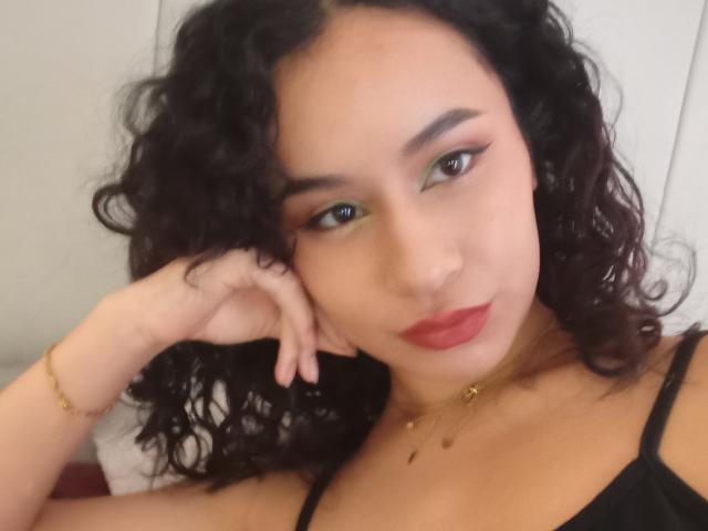 HeilyPeterson a les cheveux noirs bouclés, du fard à paupières vert, du rouge à lèvres, et porte un haut noir. Elle a une main posée sur sa joue et regarde l'appareil photo..