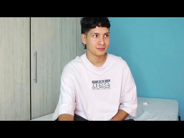 AdonissHot is a cute young dude with short black hair, wearing a white tee, sitting on a bed with a blue wall and wooden closet in the background.