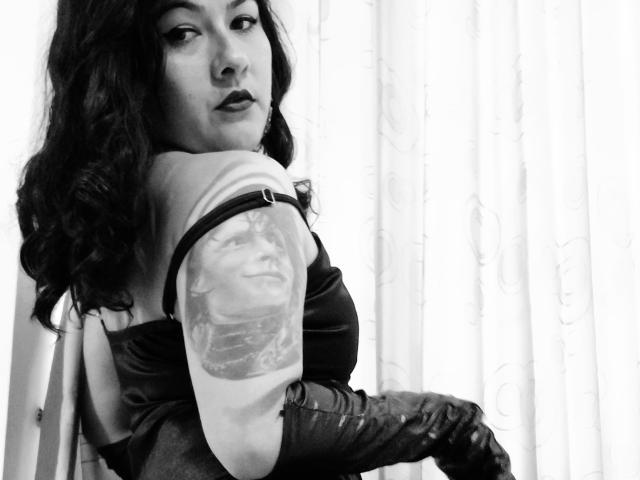 FreeMystic69, avec ses cheveux noirs ondulés, son rouge à lèvres foncé, ses gants en cuir et son tatouage sur le bras, regardant par-dessus son épaule, photo en noir et blanc. Seins, tatouage sur le bras, sexy, confiante, séduisante..