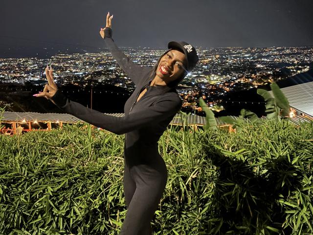 SherilRuiz, wearing a black suit and cap, stands with arms raised, smiling, in a green bamboo field at night, city lights in the background. Her ass is not visible.