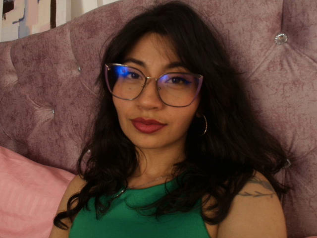 Selene Leon, com cabelos escuros e ondulados, vestindo uma blusa verde, óculos com um leve tom de roxo, batom vermelho e brincos de argola, tem uma tatuagem no ombro direito. Ela está sentada em uma cama rosa..