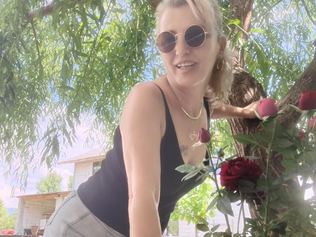 SexyCoco con i capelli biondi, che indossa occhiali da sole rotondi, una canotta nera, pantaloni grigi, una collana dorata, appoggiata a un albero, con una rosa in mano, sorridente, in un ambiente esterno soleggiato, con rami di alberi sullo sfondo..