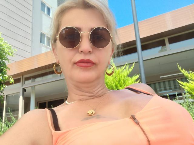SexyCoco has blonde hair, round sunglasses, gold hoop earrings, red lipstick, a peach tank top showing her tits, and a small tattoo below her collarbone.