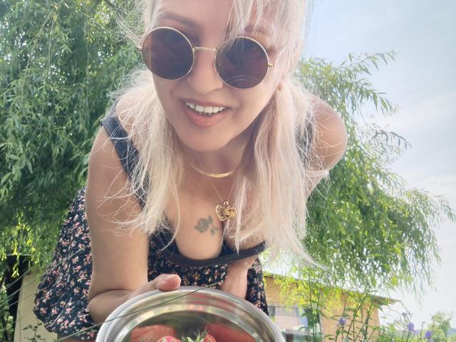 SexyCoco with blonde hair, wearing round sunglasses and a floral dress, leaning forward with a tray of red tomatoes, showing small tits and a tattoo under her cleavage.