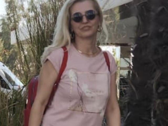 SexyCoco, com cabelo loiro, usando óculos de sol escuros, uma camiseta rosa com uma estampa, uma mochila vermelha e um colar, parada do lado de fora com uma palmeira ao fundo..