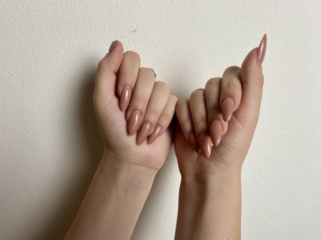 Les mains de MarriAnni, avec de longs ongles roses en forme d'amande, les doigts légèrement recourbés, contre un mur blanc. Son annulaire porte une petite bague discrète..