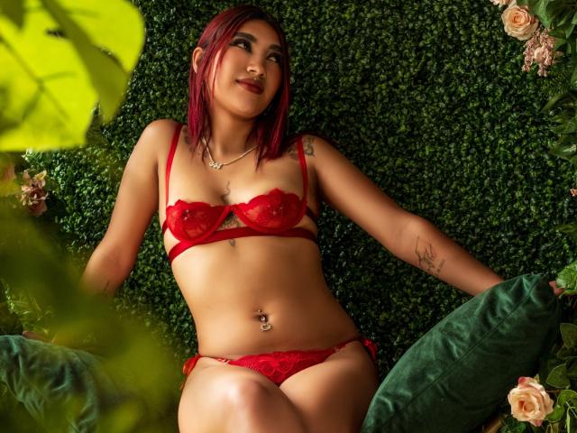 NemesisSinner con reggiseno e slip rossi, capelli rossi, tatuaggi, piercing all'ombelico, sdraiata su un cuscino verde, circondata da rose, foglie verdi in primo piano, che guarda verso l'alto con un leggero sorriso..