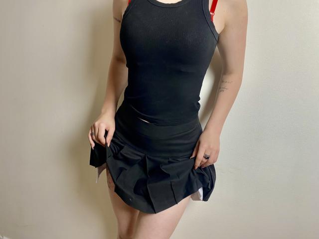 MarriAnni's black tank top and short black skirt show her pale skin and red straps, with her hands holding the skirt's sides, revealing her bare thighs and tattoos.