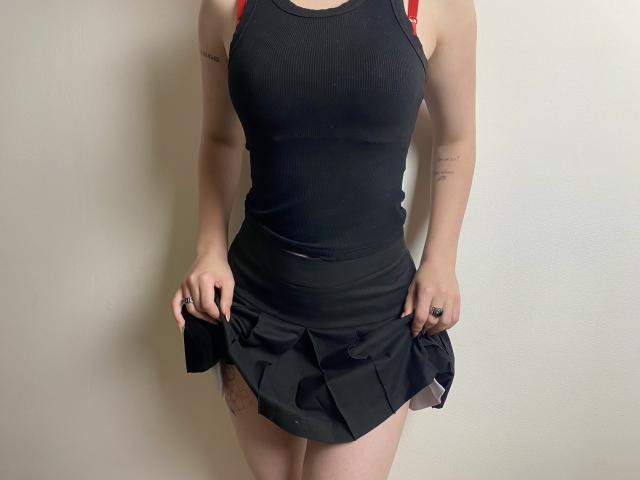 MarriAnni's black tank top, showing her tits, with a red strap visible. She's pulling up a black skirt, revealing a tattoo on her right thigh. White background.