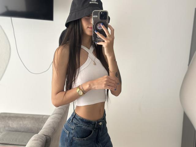 CiaSweet with long dark hair, wearing a black bucket hat, white crop top, and blue jeans, holding a phone, showing a slim waist and small tits, in a mirror selfie.