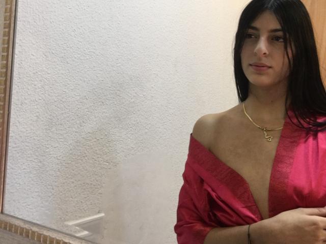 CiaSweet with long black hair, red off-shoulder top, showing cleavage, gold necklace, standing, looking at camera, slightly smiling, right arm crossed.