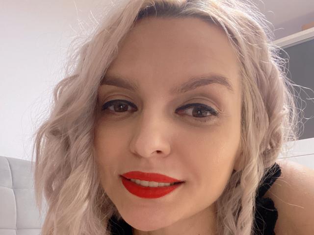 DouceAmelia has wavy, platinum blonde hair, red lipstick, and black eyeliner, smiling with brown eyes, and wearing a black top, in a selfie.