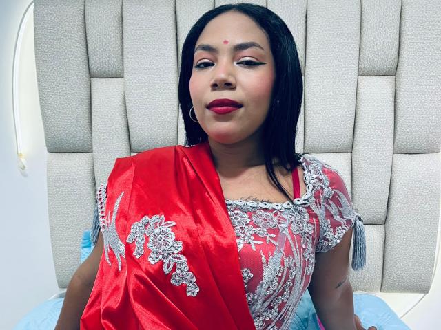 KalindaSajanii, with red sari and silver floral embroidery, sitting on bed, red lipstick, black hair, red bindi on forehead, smiling, gray padded headboard in background.