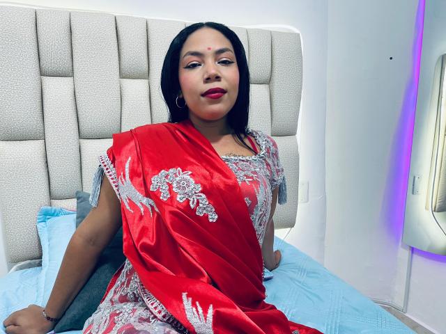 KalindaSajanii with medium brown skin, black hair, and red eyes, wearing a shiny red sari with silver embroidery, sitting on a bed with a blue pillow.