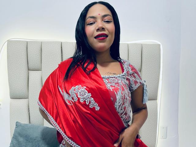 KalindaSajanii with red saree, silver embroidery, and red lipstick, smiling, black hair, medium tits, hand on hip, white headboard, gray pillow, and subtle tattoo on left arm.