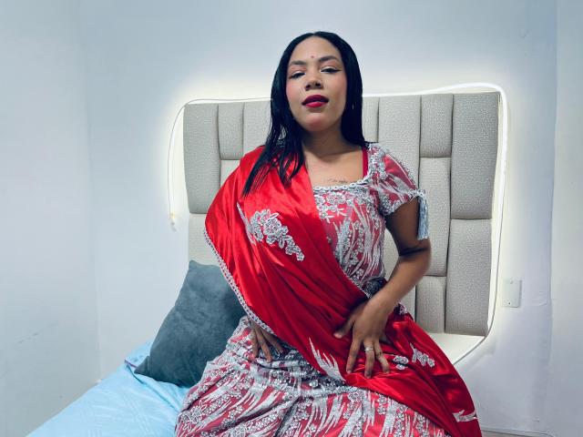 KalindaSajanii, with medium brown skin, black hair, and red eyes, wears a red and white floral dress, sitting on a bed, showing her ass, with a confident smile.