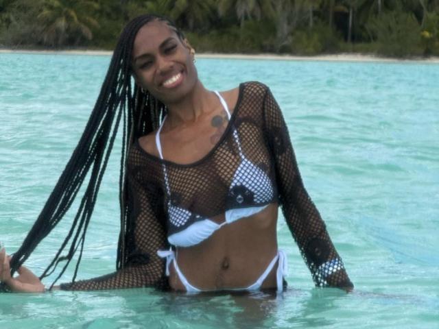 SherilRuiz with dark skin and long braids, wearing a mesh top over a white and black checkered bikini, smiling in clear blue water, showing her small tits.