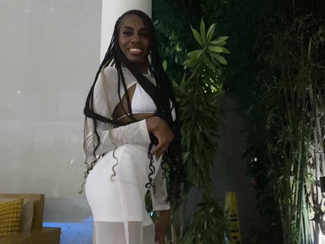 SherilRuiz with long black braids, wearing a white crop top and tight white pants, smiling with her hand on her butt, standing next to a green plant.