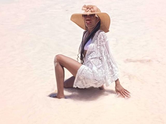 SherilRuiz with long braids, wearing a white lace dress, wide-brimmed straw hat, and sunglasses, sitting in pale sandy beach, her legs slightly apart, showing her bare ass.