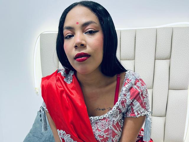 KalindaSajanii with long black hair, red lipstick, and a red saree with silver lace. She's got a red bindi on her forehead and a tattoo on her chest.