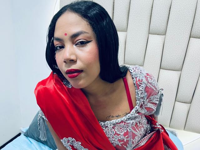KalindaSajanii with red sari, white lace, and red lipstick, black bob, red dot on forehead, sitting on bed, showing small tattoo on chest, looking at camera.