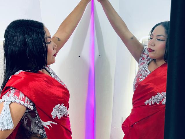 KalindaSajanii with long black hair, red sari with white embroidery, arms raised, reflecting in mirror, tattoo on right arm, purple light in background, serious expression, red lipstick.