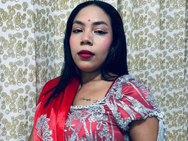 KalindaSajanii with dark hair, red lipstick, and a red and silver floral dress, featuring a bindi on her forehead and a tattoo under her chin, looking at the camera.