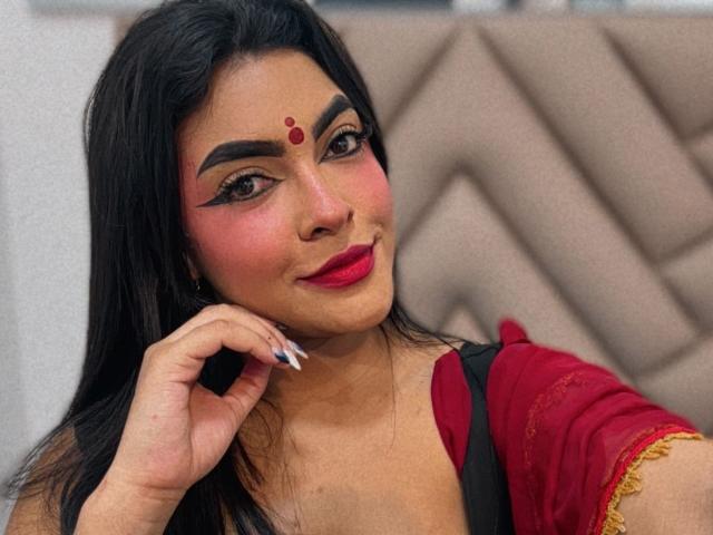 LakshmIrani has dark hair, red lipstick, bold eyeliner, and a red dot on her forehead. She's wearing a red top with black and gold details. Her nails are long.