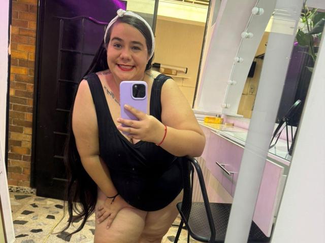 AndreaaX69, formosa con lunghi capelli neri, sorridente, indossa un vestito nero, mostra il sedere, tiene in mano un telefono viola, è in piedi di fronte a uno specchio, con un muro di mattoni sullo sfondo..