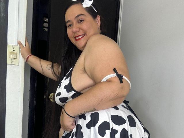 AndreaaX69, formosa con grandi seni, indossa un vestito a pois di mucca, una fascia per capelli bianca con un fiocco, lunghi capelli neri, rossetto rosso, tatuaggio sul braccio, in piedi davanti a una porta, sorridente, braccio destro alzato..