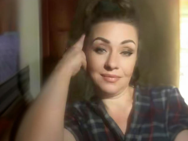 DinaraQMoon with brown hair in a ponytail, wearing a plaid shirt, has a hand on her cheek, smiling with subtle makeup, showing her soft-looking tits.