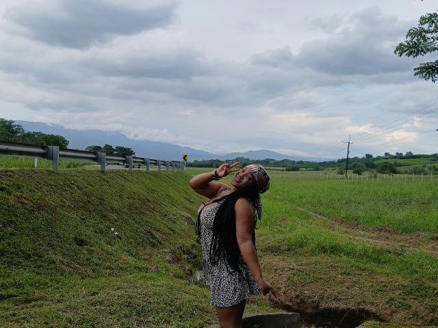 DakotaScoth with dark skin and long black hair, wearing a sleeveless, patterned dress, drinking from a bottle, standing on grassy slope, mountains in background, cloudy sky.