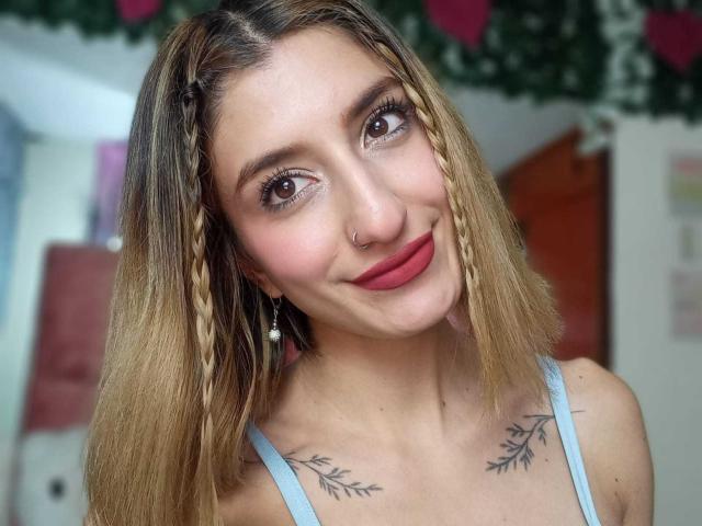 Kaiix a les cheveux blonds tressés, du rouge à lèvres, un anneau au nez et des tatouages sur la poitrine. Elle sourit, porte un débardeur bleu, a les yeux marron et porte des boucles d'oreilles de perles..