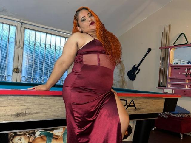 CoolSweets with long red hair in a sheer, burgundy dress, showing her ass with a high slit, leaning on a pool table, looking confident.