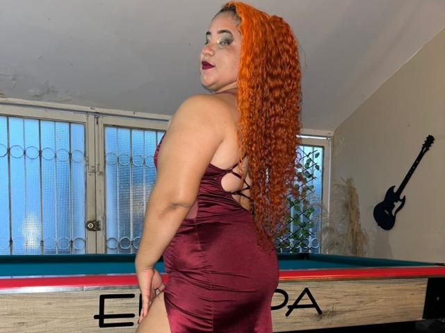 CoolSweets with curly orange hair, wearing a tight burgundy dress, showing her ass, standing by a pool table, looking back with dark lipstick.